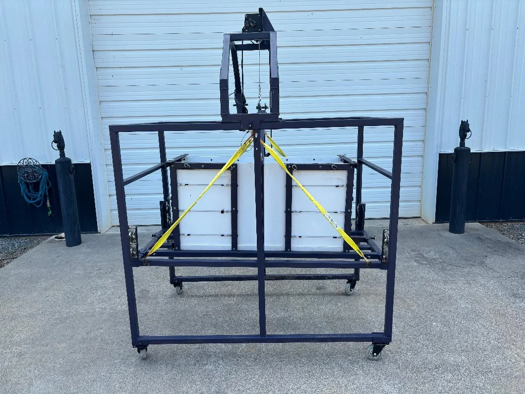 The Juniper Pig Lift — a dark steel wheeled frame with an overhead winch and yellow safety webbing supporting a padded central bar.