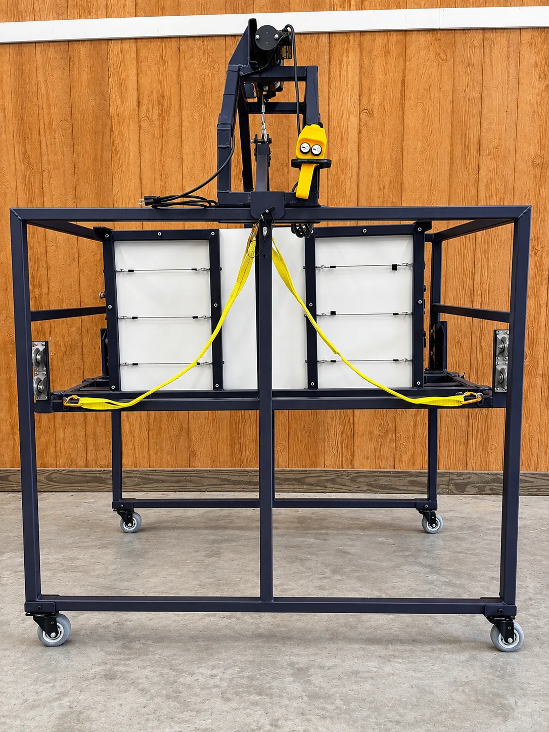 Front view of the Juniper Pig Lift — a dark steel four-caster frame with the winch arm overhead, yellow pendant control, and webbing cradling the raised chute.