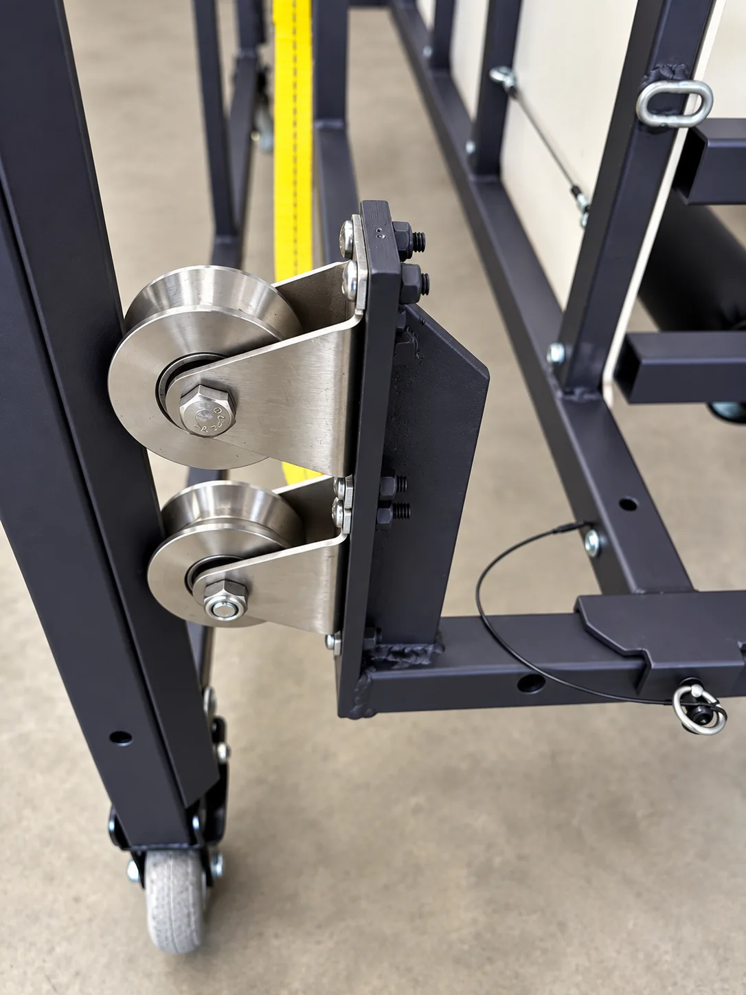 Close-up of two stainless steel guidewheels bolted to the frame, routing the yellow lift webbing, with a caster wheel below.